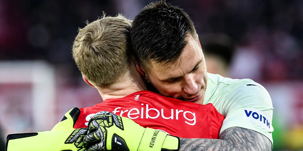 Zwei Spieler, einer vom SC Freiburg im roten Trikot und einer vom 1. FC Heidenheim im hellen Torwarttrikot, umarmen sich nach dem Spiel auf dem Platz. (Foto)