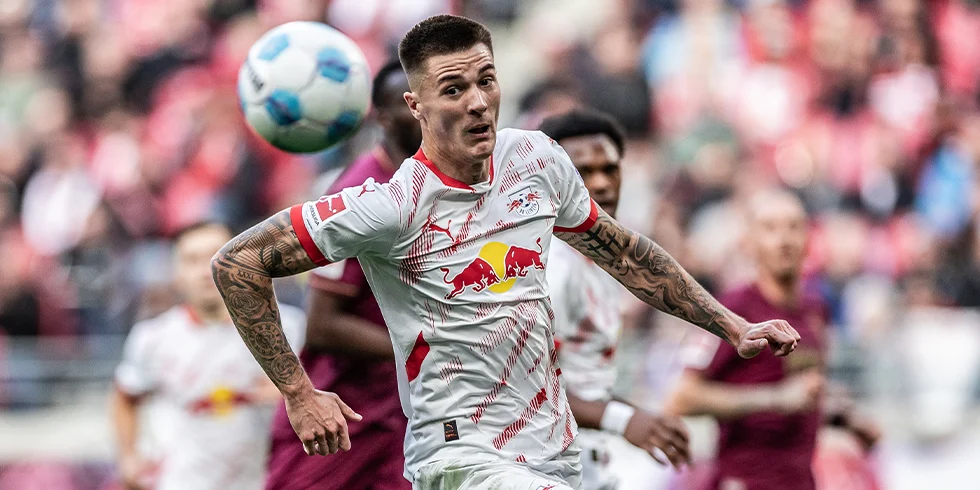 Ein RB Leipzig Spieler im weißen Trikot mit dem roten Bullen-Logo läuft dem Ball entgegen, während Gegenspieler in dunkelroten Trikots im Hintergrund zu sehen sind. (Foto)
