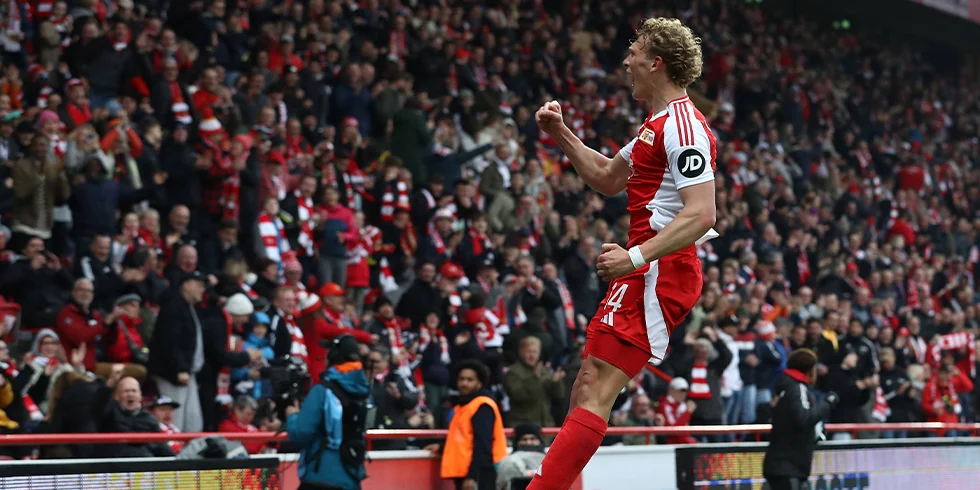 Leopold Querfeld im roten Union Berlin Trikot springt jubelnd in die Luft – fast so, als würde er gleich abheben wie ein Superheld vor der jubelnden Fankurve. (Foto)