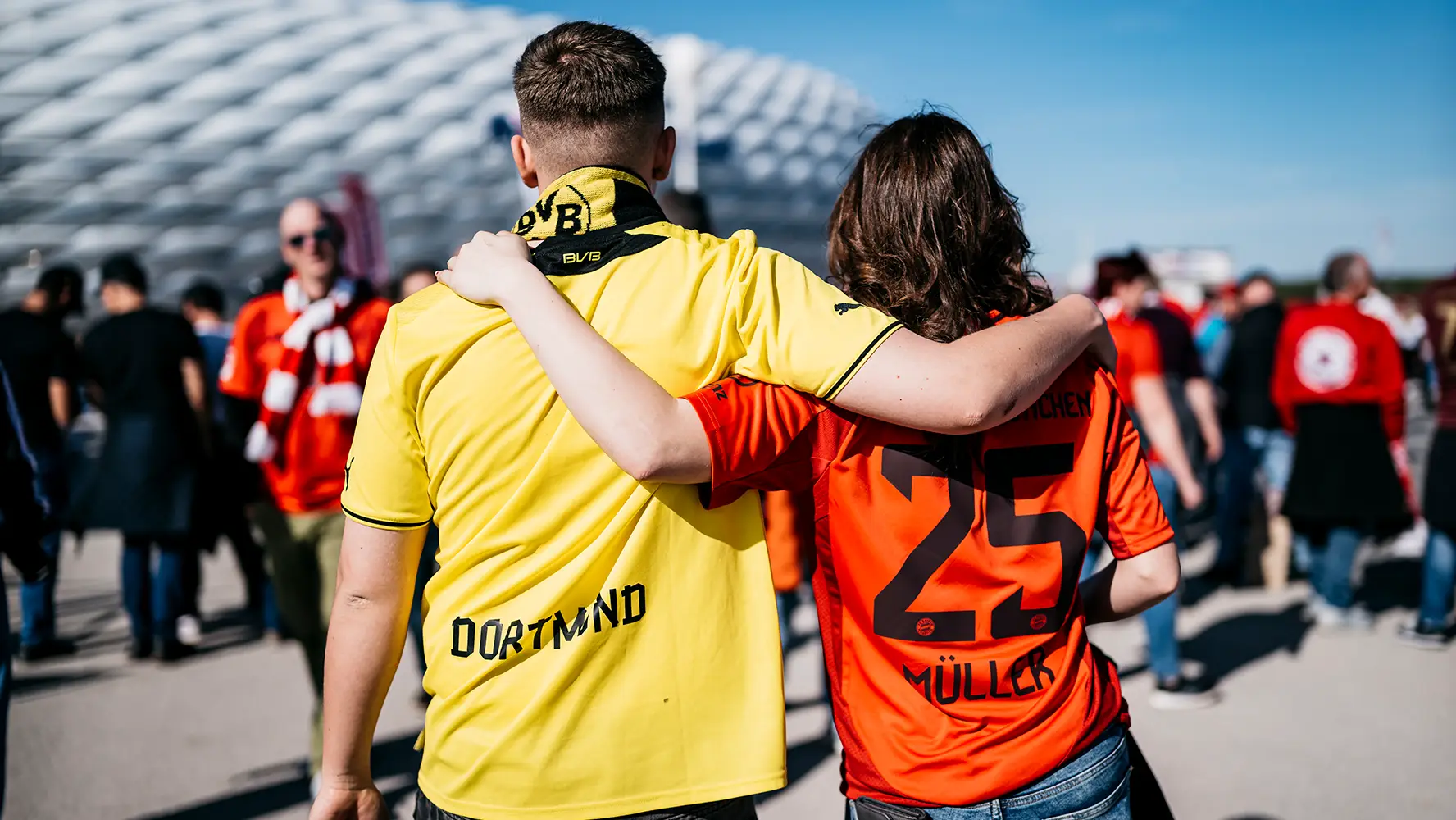 Ein Mann in BVB Trikot und eine Frau in FC Bayern Trikot stehen vor einem Stadion. Sie legen die Arme um die Schultern des anderen. (Foto)